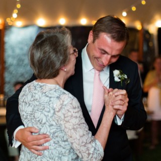 Groom shares an emotional mother-son dance under glowing string lights at a seaside wedding.