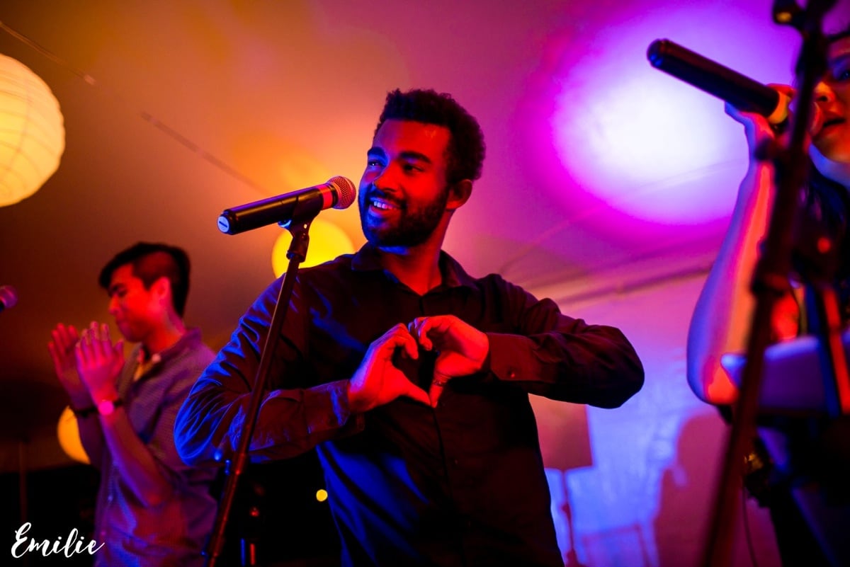 Singer making a heart gesture on stage under vibrant purple and yellow lights during live performance.