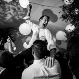 Groom lifted joyfully by friends under glowing lanterns at lively Bar Harbor wedding celebration.