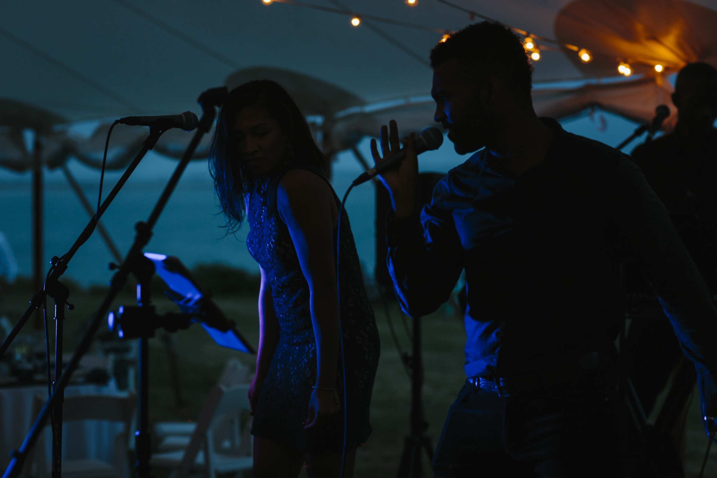Evening live music duo performing under romantic string lights at a waterfront wedding reception.