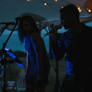 Evening live music duo performing under romantic string lights at a waterfront wedding reception.