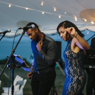 Male and female vocalists perform under glowing string lights at a seaside wedding celebration.