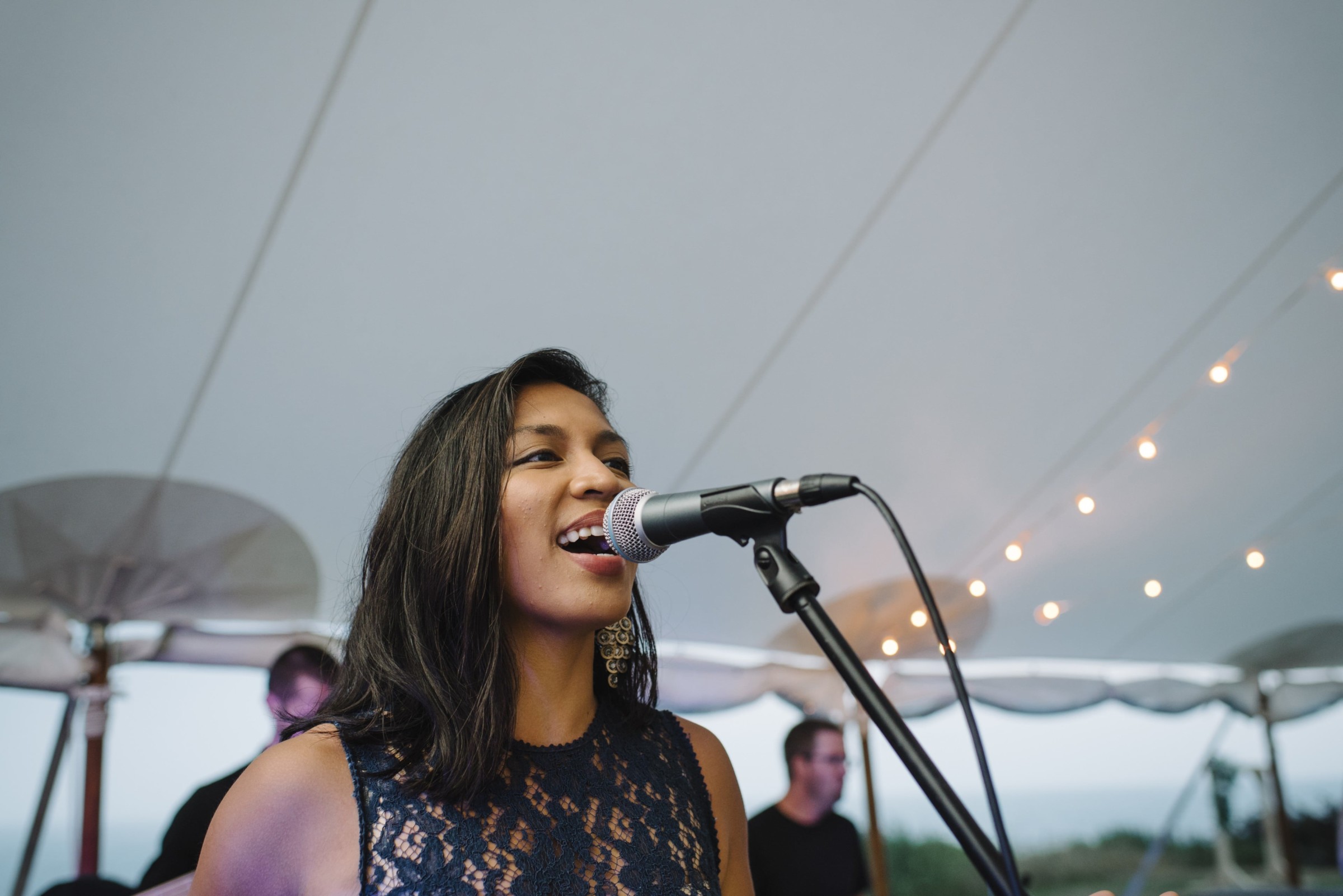 Woman singing passionately at outdoor wedding under canopy with live band and warm string lights.
