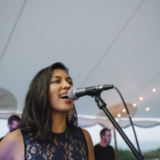 Woman singing passionately at outdoor wedding under canopy with live band and warm string lights.