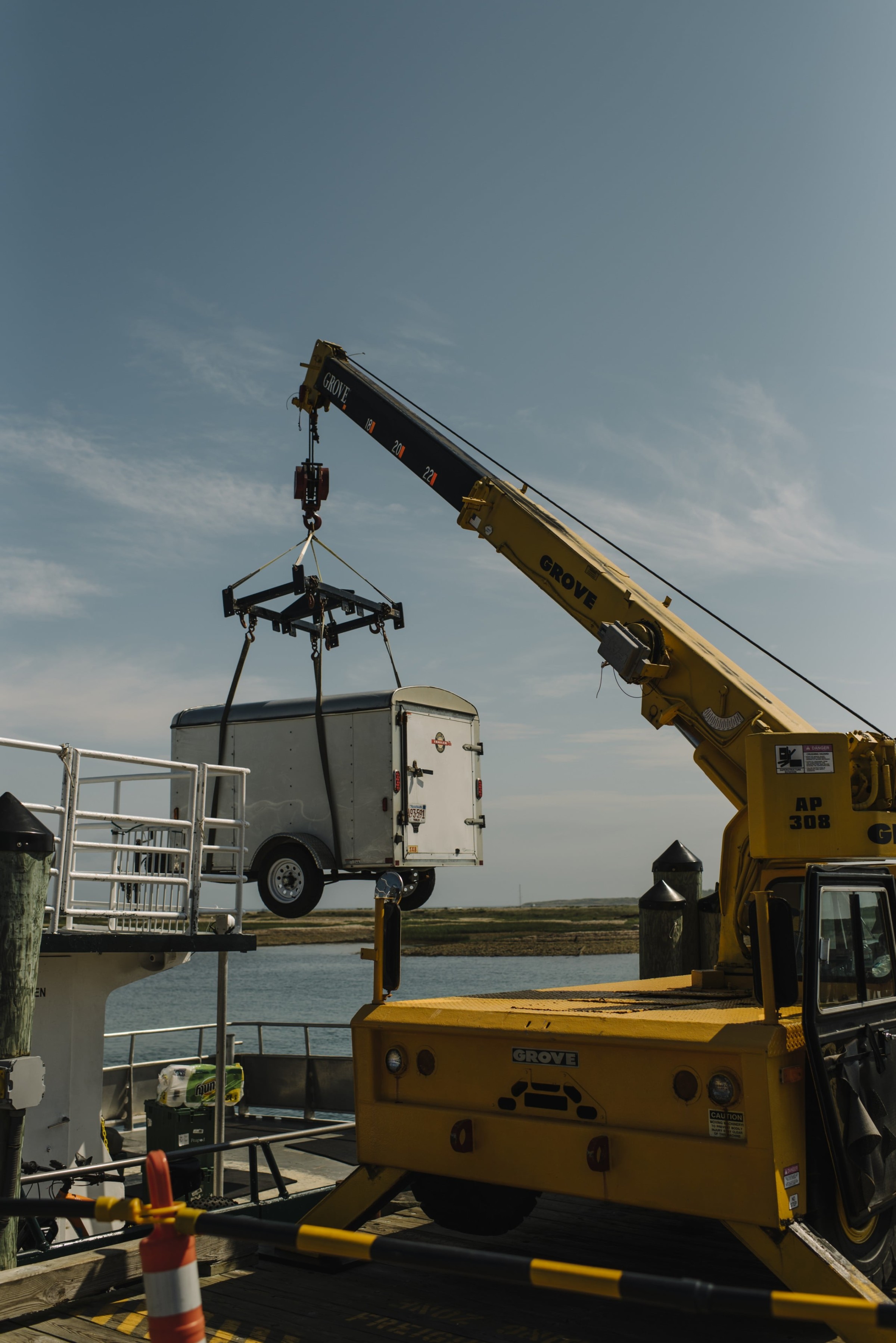 Yellow Grove crane lifting white cargo trailer over waterfront dock under clear blue coastal sky.