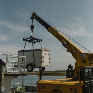 Yellow Grove crane lifting white cargo trailer over waterfront dock under clear blue coastal sky.