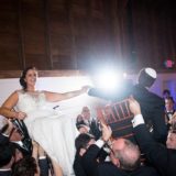 Bride and groom lifted on chairs during joyful Jewish Hora dance at Gedney Farm wedding.