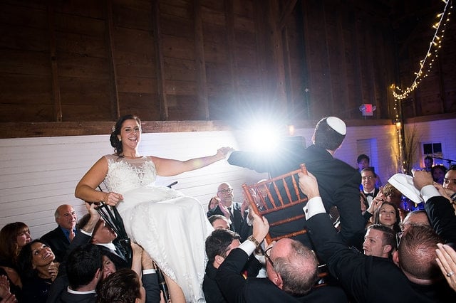 Bride and groom lifted on chairs during joyful Jewish Hora dance at Gedney Farm wedding.