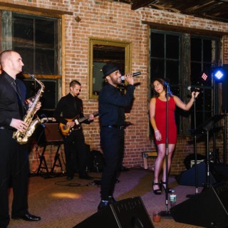 Live band performing in a rustic loft venue with brick walls and a singer in a red dress.