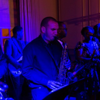 Live jazz band performing indoors under vibrant blue and purple stage lighting.