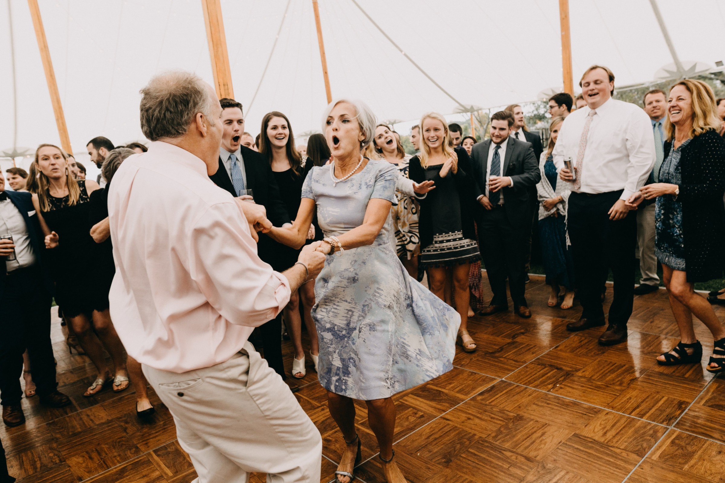 Older couple dancing joyfully at outdoor wedding reception under white tent surrounded by cheering guests.