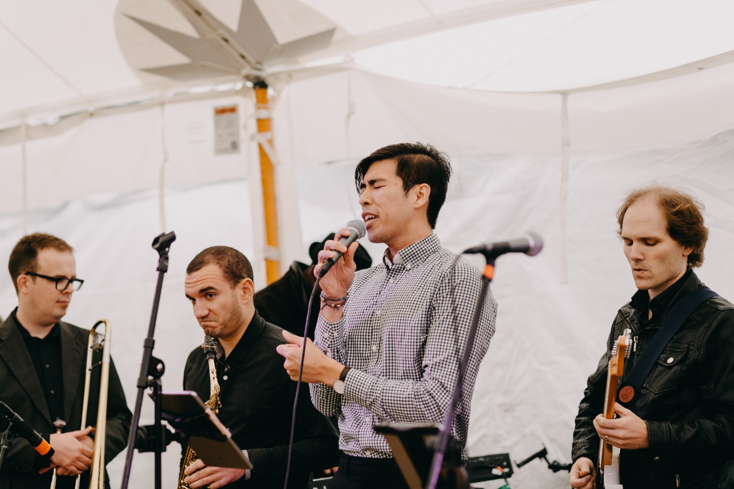 Live band performing under white tent with singer, guitarist, trombone, and saxophone players.