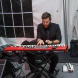 Bearded musician passionately playing red Nord keyboard under white tent at intimate live performance.