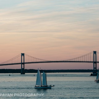 Sailboats at sunset beneath Newport Bridge reflecting pastel skies over tranquil Rhode Island waters.