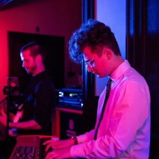 Keyboardist and guitarist performing under vibrant pink, blue, and purple stage lights at a Boston venue.