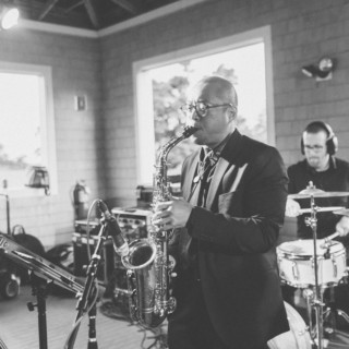 Black and white jazz duo performing indoors with saxophonist and drummer in natural light.
