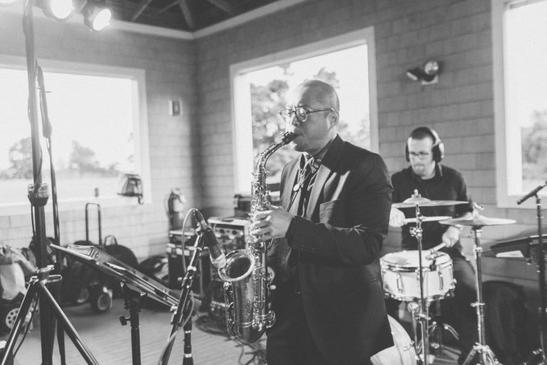 Black and white jazz duo performing indoors with saxophonist and drummer in natural light.