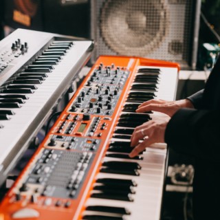 Musician playing bright orange synthesizer and white keyboard in a professional studio setup.