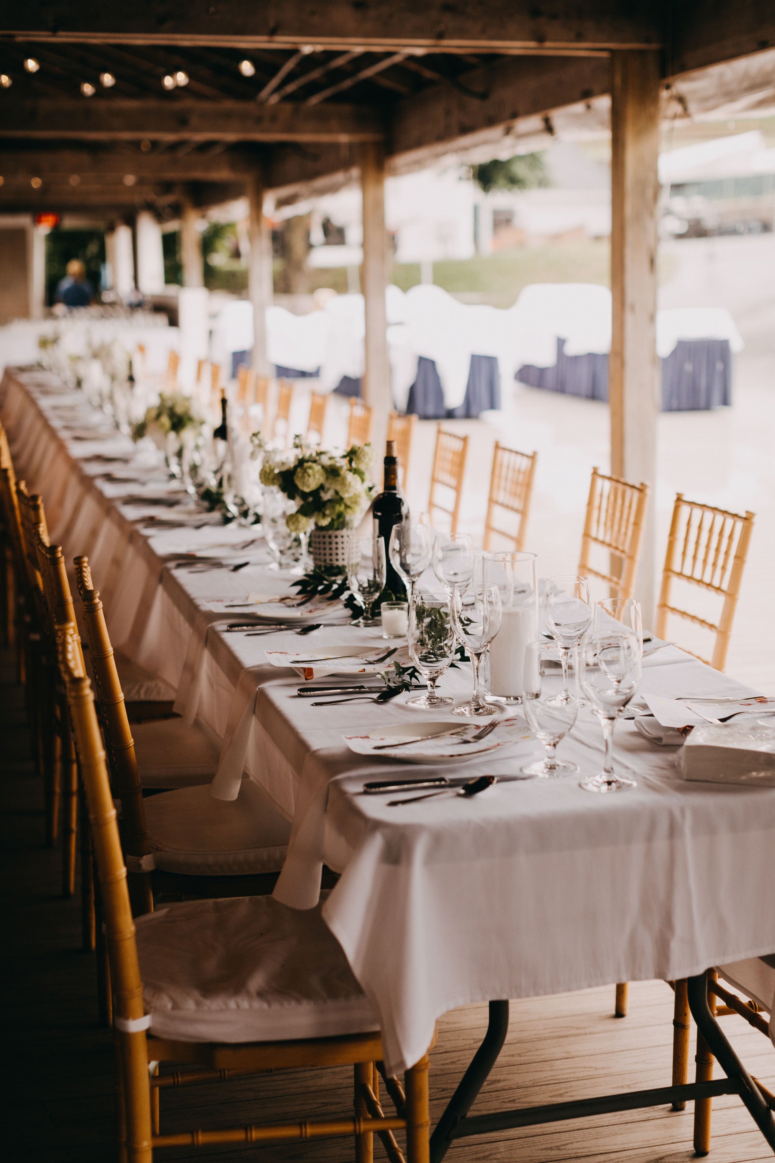 Elegant rustic wedding table with white linens, gold chairs, and floral centerpieces under wooden pavilion.