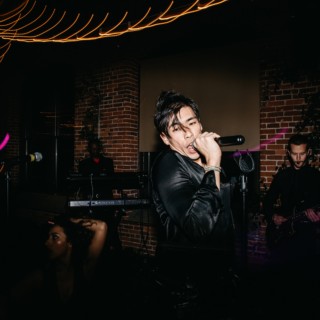 Passionate male singer performing live with band under vibrant orange and purple stage lighting.