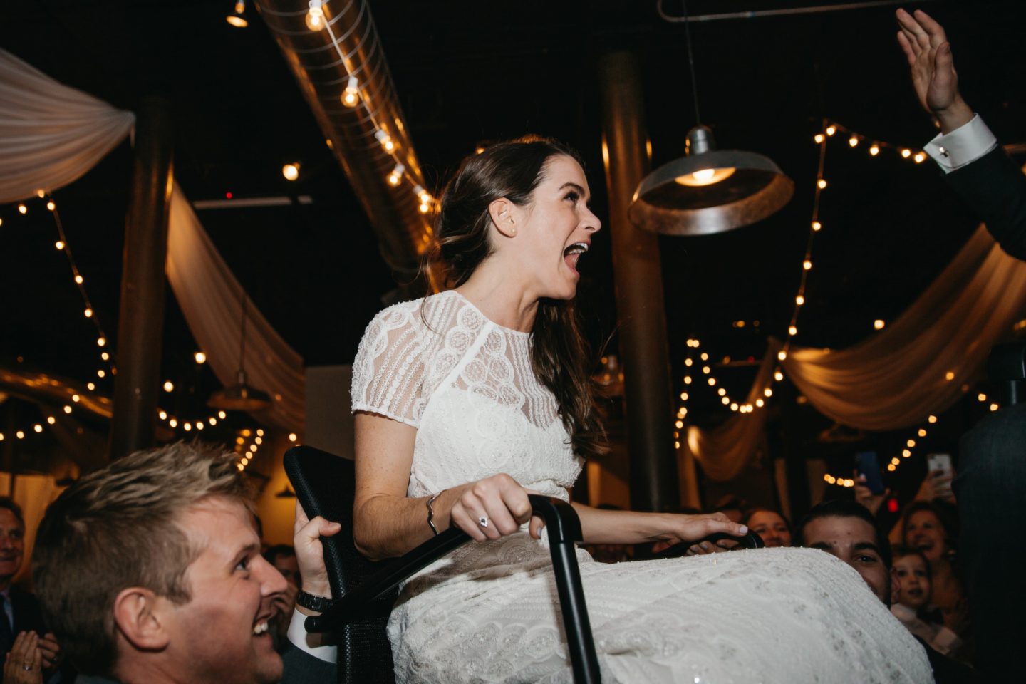 Bride lifted on chair during joyful Rivermill wedding celebration with guests and warm string lights.