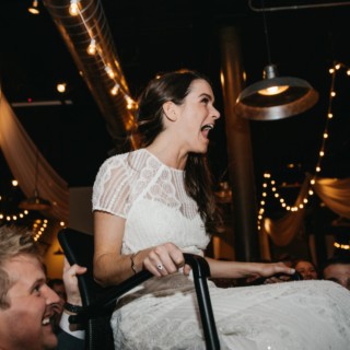 Bride lifted on chair during joyful Rivermill wedding celebration with guests and warm string lights.