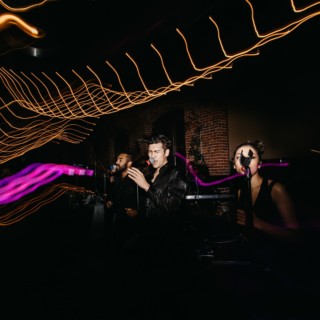 Three vocalists performing live under vibrant stage lighting with swirling golden and purple light trails.