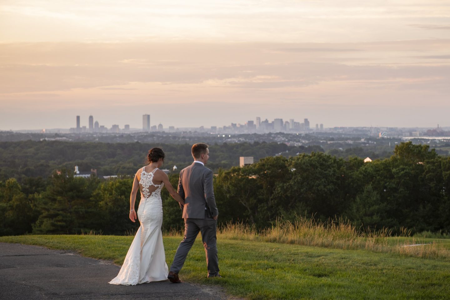 Bride and groom walking hand in hand at sunset overlooking Boston skyline at Granite Links.