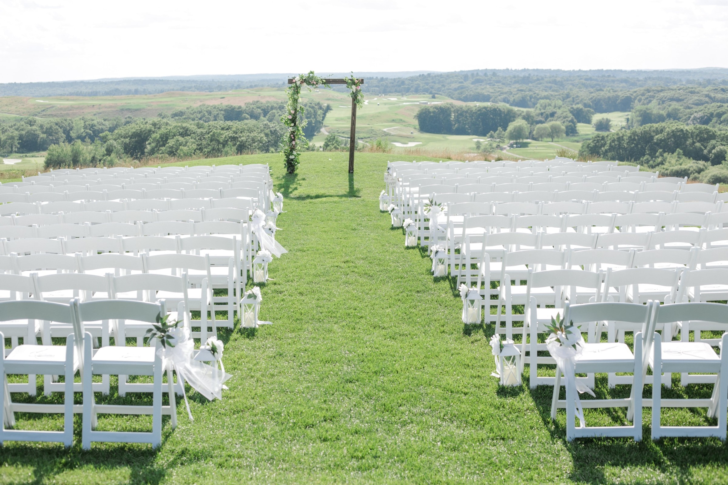 Outdoor wedding ceremony with white chairs, floral arch, and scenic hillside backdrop in Quincy, MA.