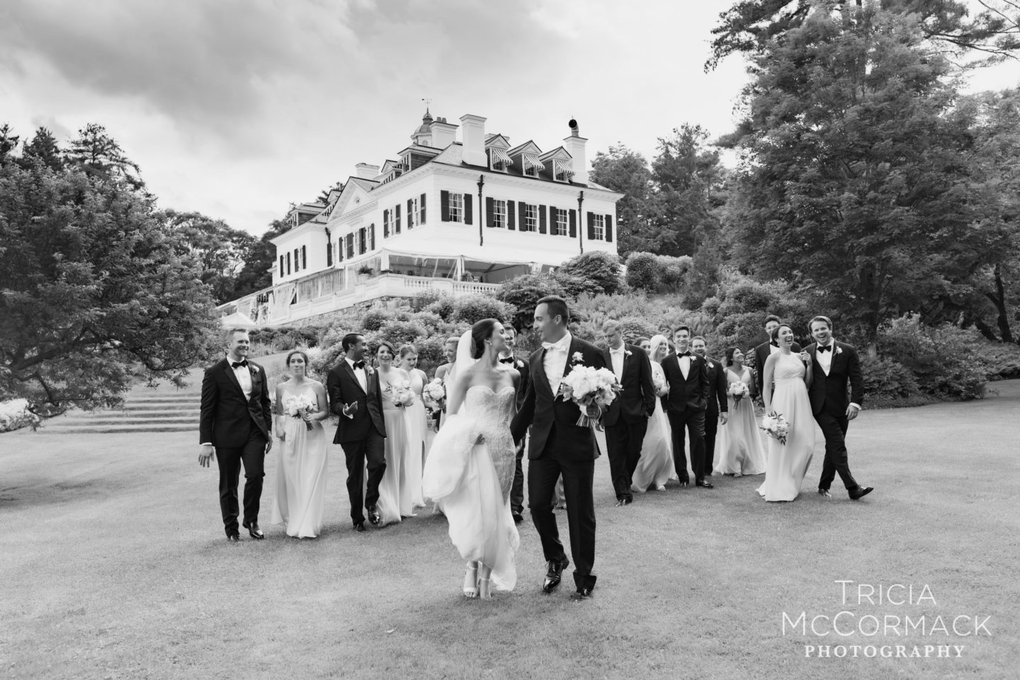 Bride and groom lead joyful wedding party across historic Lenox estate lawn in timeless black and white.