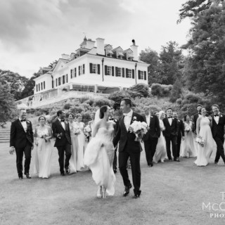 Bride and groom lead joyful wedding party across historic Lenox estate lawn in timeless black and white.