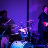 Live jazz duo performing at Newport Marriott with drummer and guitarist under purple lighting.