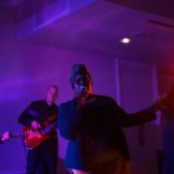 Female singer performing live with band under vibrant orange, pink, and blue stage lighting.