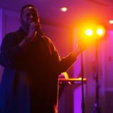 Man singing passionately on stage under warm lights with colorful blue and violet background.