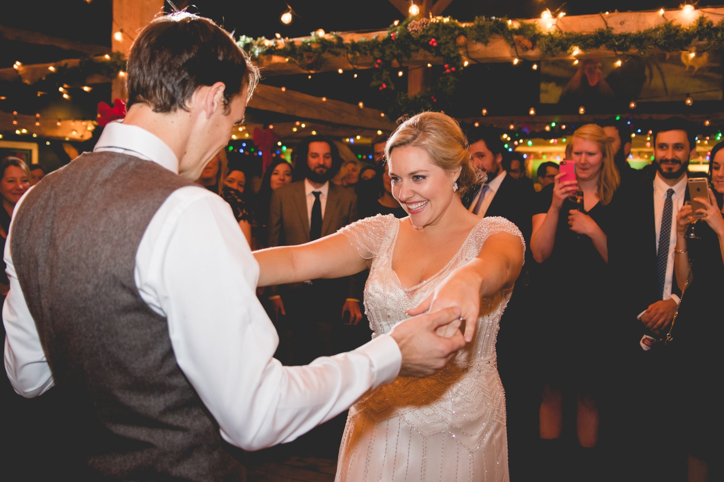 Bride and groom share first dance at rustic winter wedding surrounded by guests and warm lights.