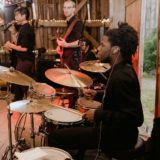 Live band performing in rustic barn with drummer, vocalist, and bassist under warm string lights.