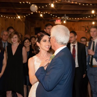 Bride and father share an emotional dance at a rustic, warmly lit wedding reception.