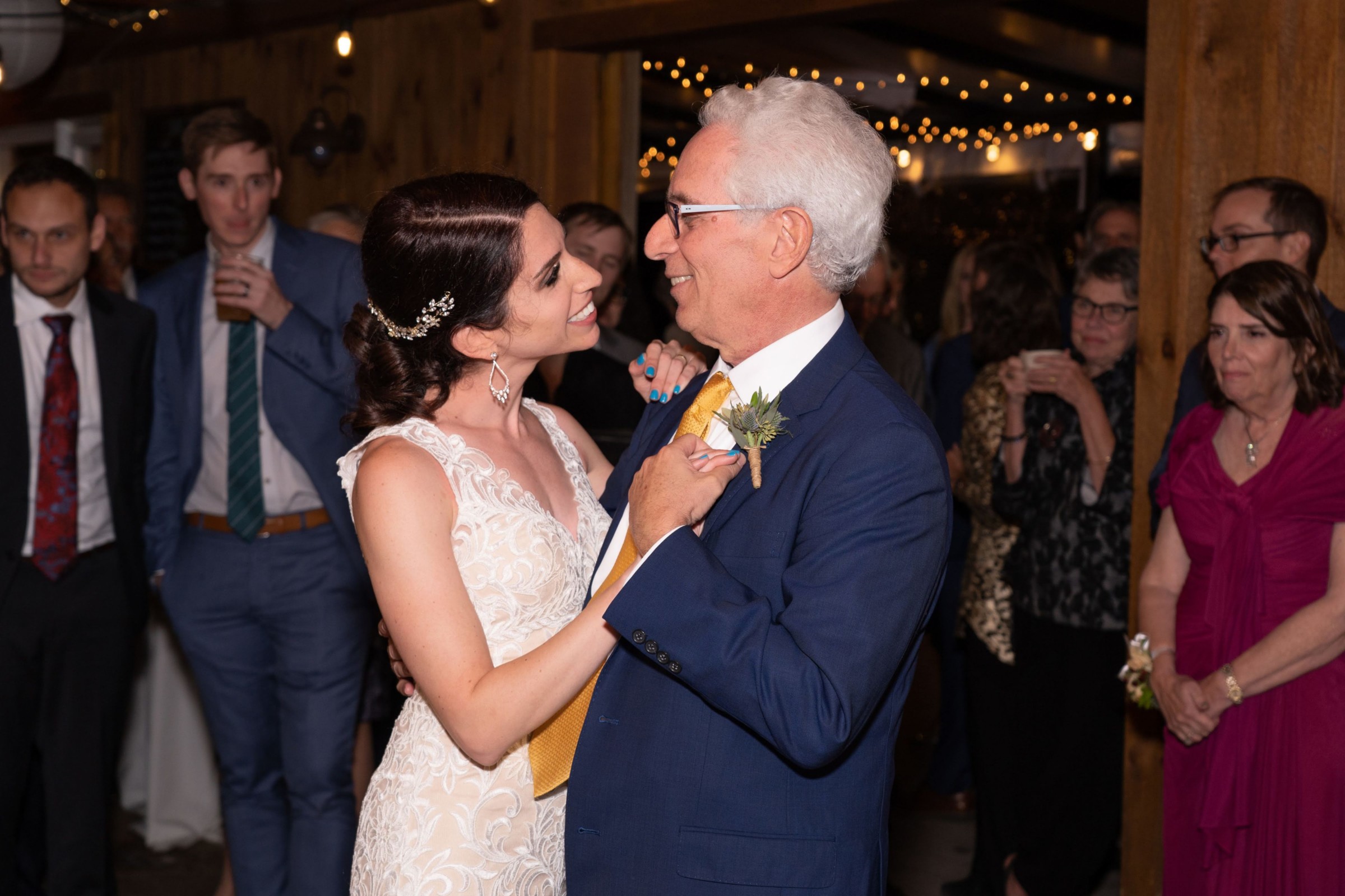 Bride shares heartfelt father-daughter dance under warm string lights at rustic wedding reception.