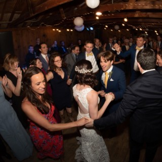 Bride and groom dancing joyfully at rustic barn wedding surrounded by cheering guests and warm lights.