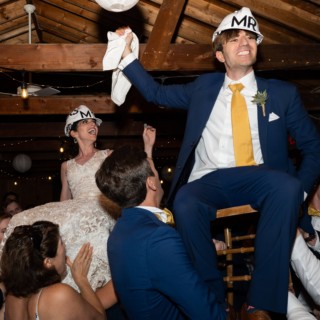 Bride and groom lifted during joyful Hora dance at rustic wedding reception.