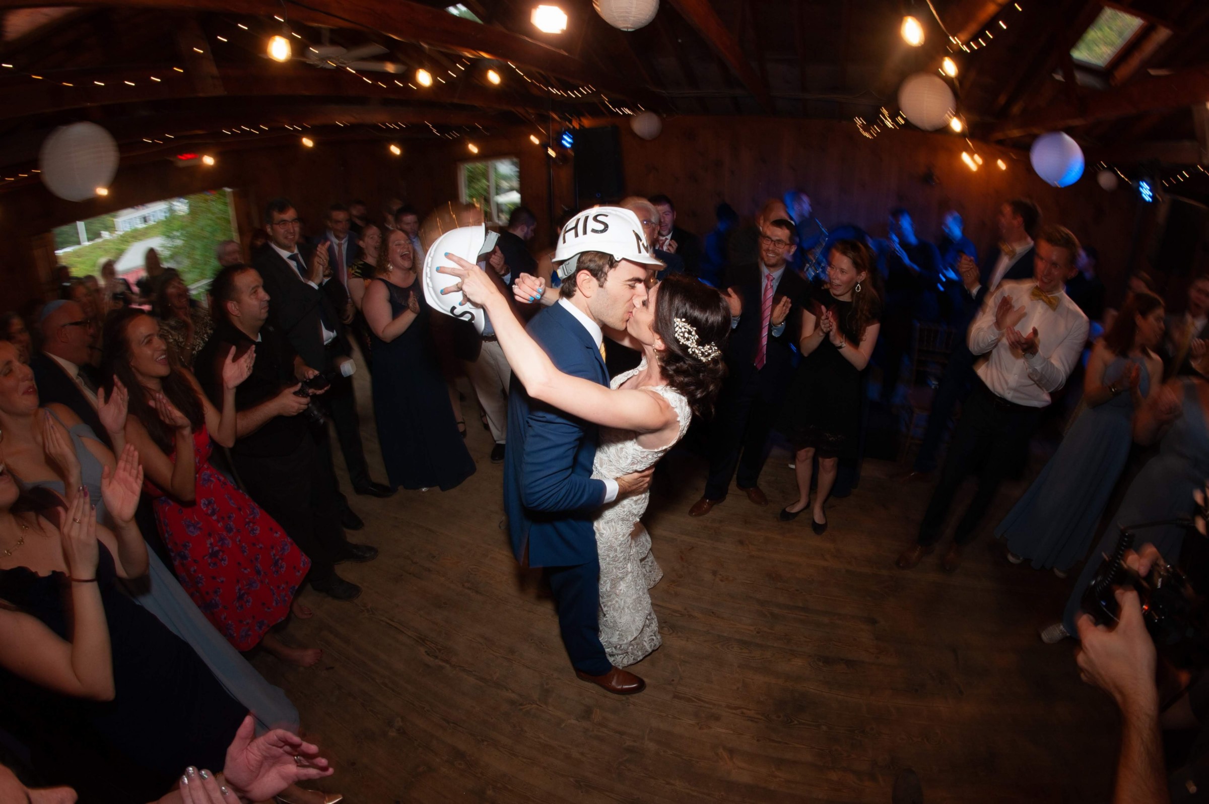 Bride and groom share a joyful kiss surrounded by cheering guests at rustic wedding reception.