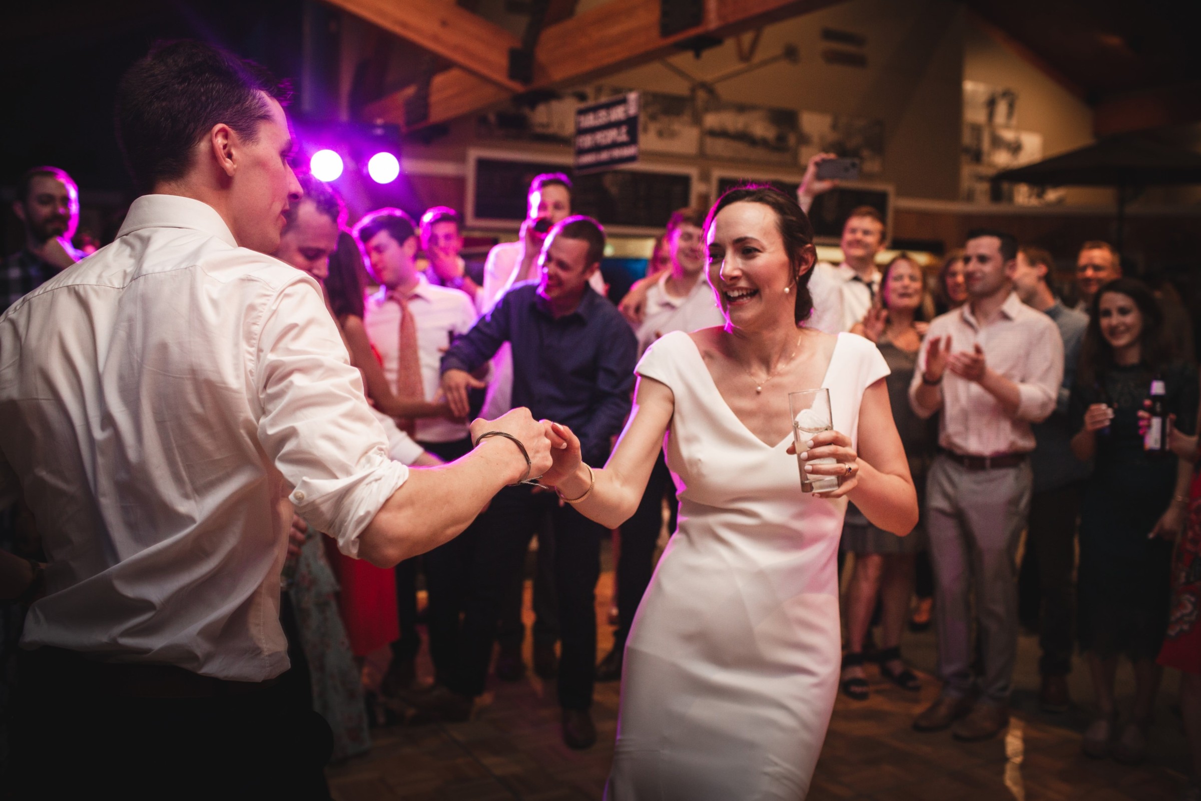 Smiling couple dancing joyfully at rustic Vermont wedding reception surrounded by cheering guests.