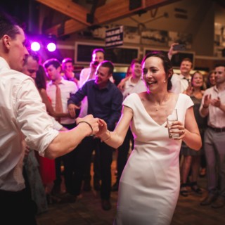 Smiling couple dancing joyfully at rustic Vermont wedding reception surrounded by cheering guests.
