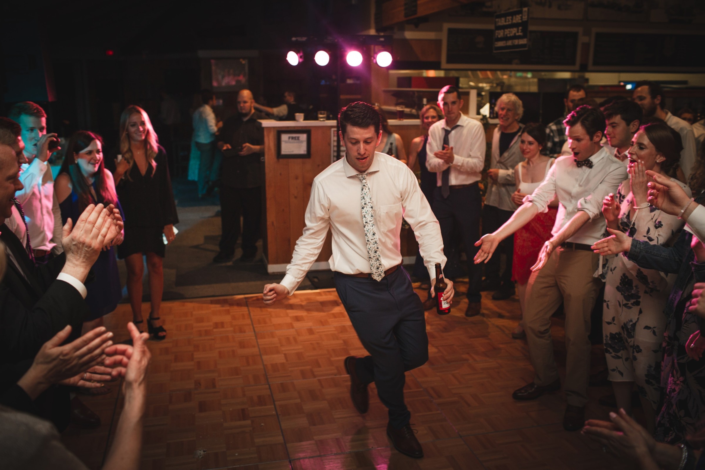 Groom dancing energetically at lively Vermont wedding reception surrounded by cheering guests and warm lights.
