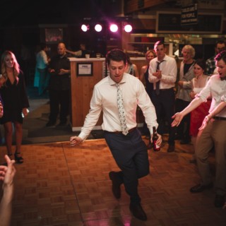 Groom dancing energetically at lively Vermont wedding reception surrounded by cheering guests and warm lights.