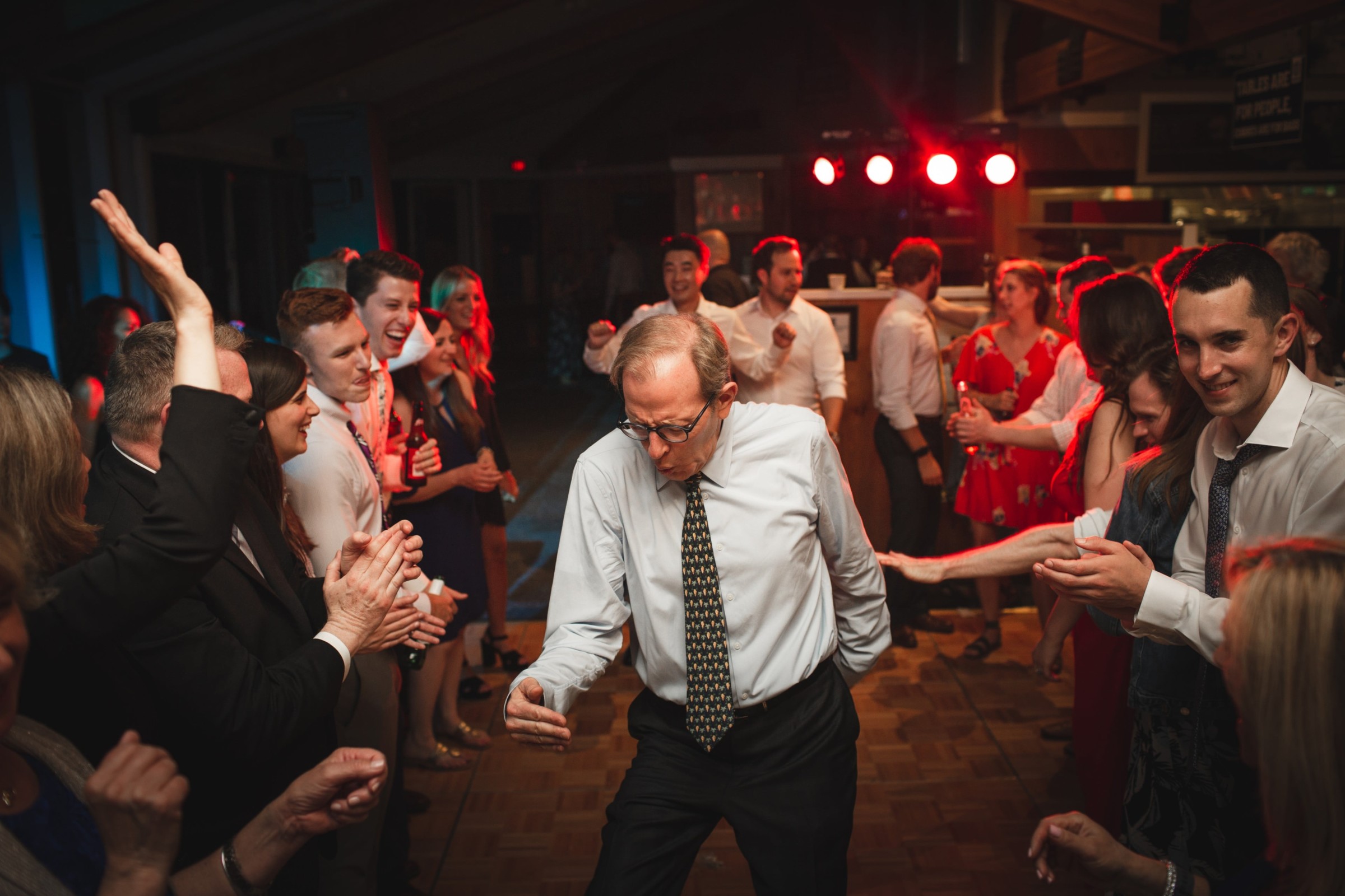 Older man dancing joyfully at Vermont wedding reception surrounded by cheering guests and warm lighting.