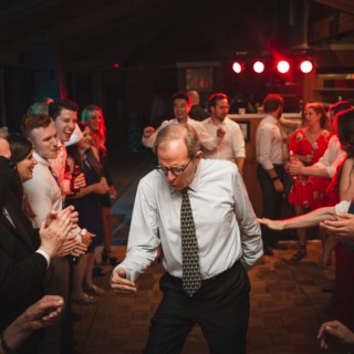 Older man dancing joyfully at Vermont wedding reception surrounded by cheering guests and warm lighting.