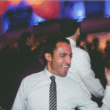 Man dancing joyfully at vibrant Boston wedding reception with colorful lights and lively crowd.