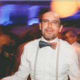 Stylish man smiling at vibrant Boston wedding with colorful lights and lively party atmosphere.