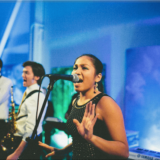 Female vocalist performing with live band under colorful stage lights at Boston event.